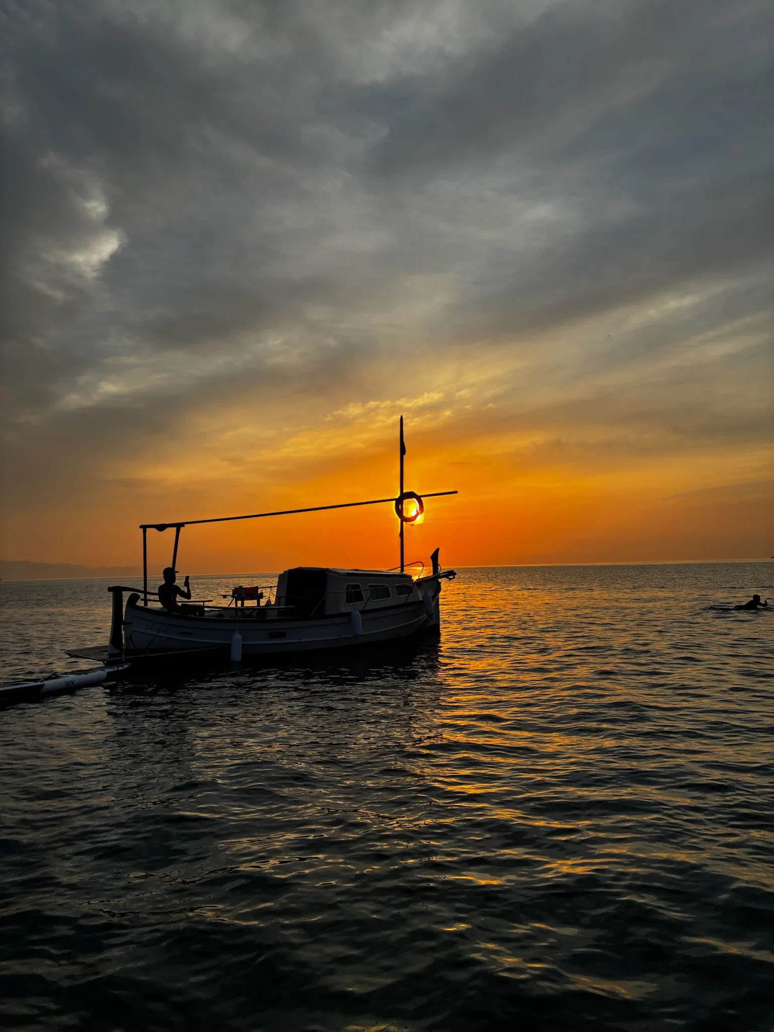 Boat at sea during sunrise, with golden reflections on the water and a person paddleboarding in the distance.